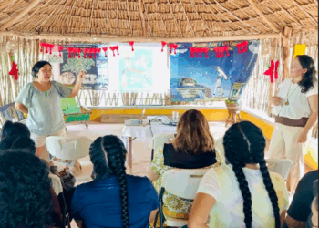 Mujeres en comunidad intercambiando saberes y experiencias educativas en un taller rural, rodeadas de niñas, niños y materiales pedagógicos.