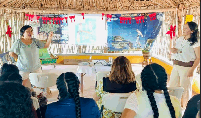 Mujeres en comunidad intercambiando saberes y experiencias educativas en un taller rural, rodeadas de niñas, niños y materiales pedagógicos.